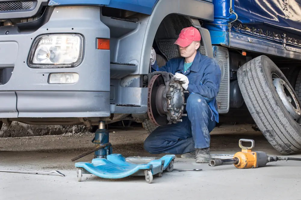 A mechanic in a red cap repairs the front wheel of a large blue truck at a busy repair shop.