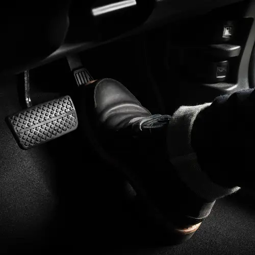 A person’s foot in a black shoe presses a car’s brake pedal, as seen inside an auto repair shop’s dim vehicle interior.