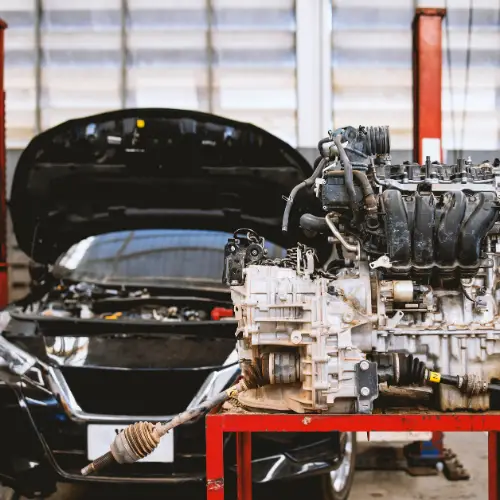 A car with its hood open in an automotive shop, engine on a red stand in front.