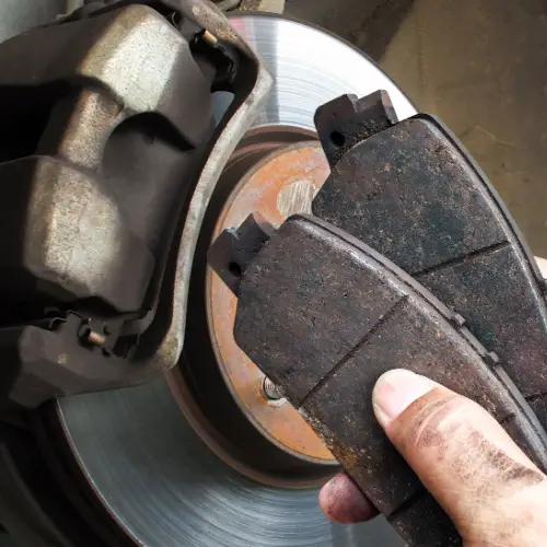 A mechanic holds two worn brake pads near a car’s brake disc and caliper, showing signs of use and grime.
