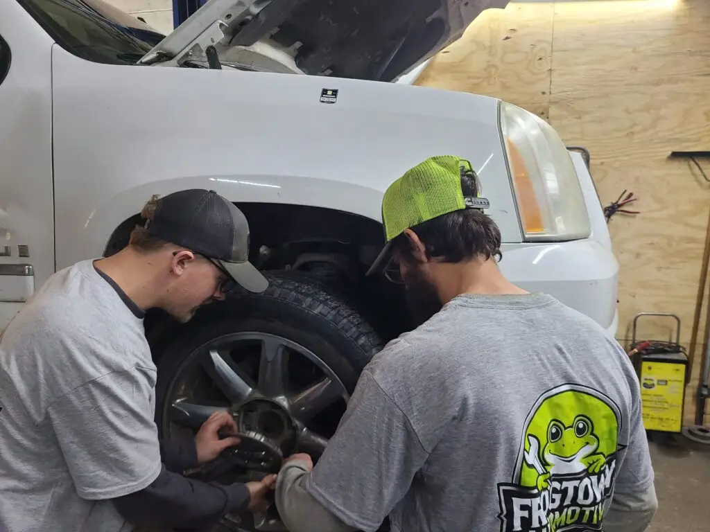 Two mechanics in hats perform auto repair on a white SUV’s wheel in a garage, with the hood raised.