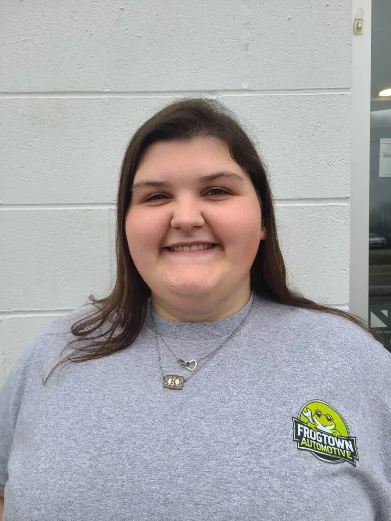 A woman with long brown hair smiles in a gray Frogtown Automotive Shop shirt, standing by a white wall.