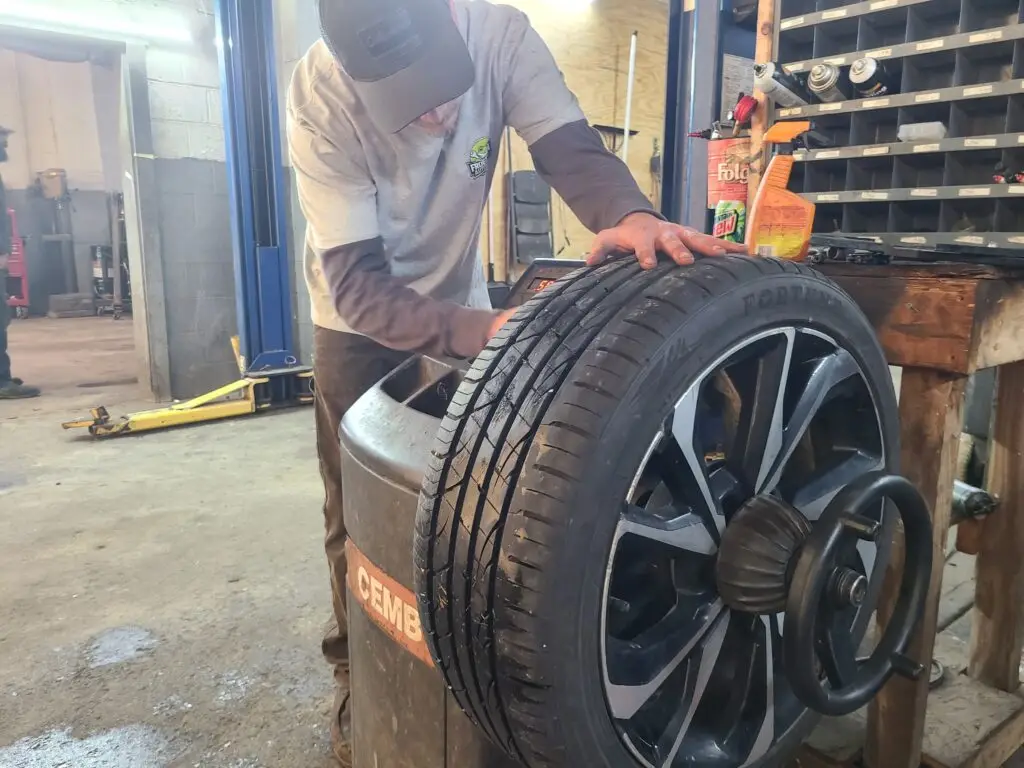A mechanic balances a car tire on a machine in an automotive shop, with tools and supplies on a shelf nearby.