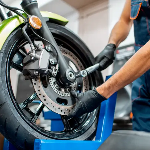 Mechanic in gloves working on a motorcycle front wheel with tools in a busy repair shop or garage setting.