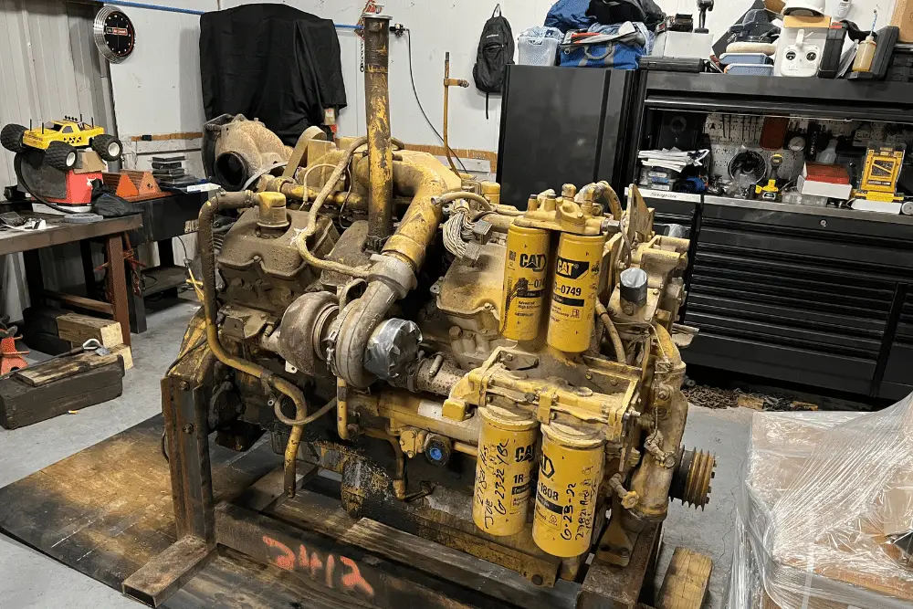 A large yellow Caterpillar diesel engine sits in an auto repair shop surrounded by tools, parts, and storage cabinets.