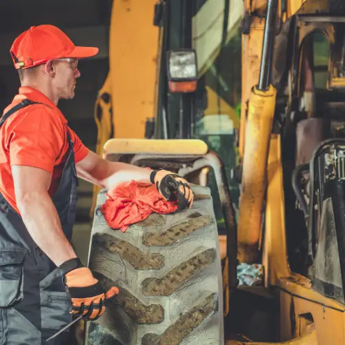 Man in workwear and gloves cleaning a large tractor tire at an auto repair shop near heavy construction equipment.