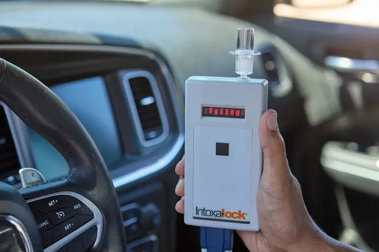 A hand holds an Intoxalock breathalyzer inside a car at a repair shop, near the steering wheel.