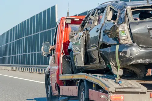 A tow truck carries a severely damaged black car to an auto repair shop on a highway next to a noise barrier wall.