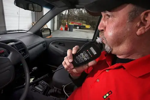 A man in a red shirt uses a breathalyzer device inside his car, possibly after visiting an automotive shop.