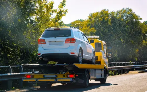 A white SUV is towed by a yellow truck to a car repair shop on a sunny day near trees and a road barrier.