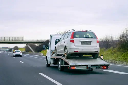 A white car from an automotive shop is transported on a flatbed tow truck along a highway under an overcast sky.