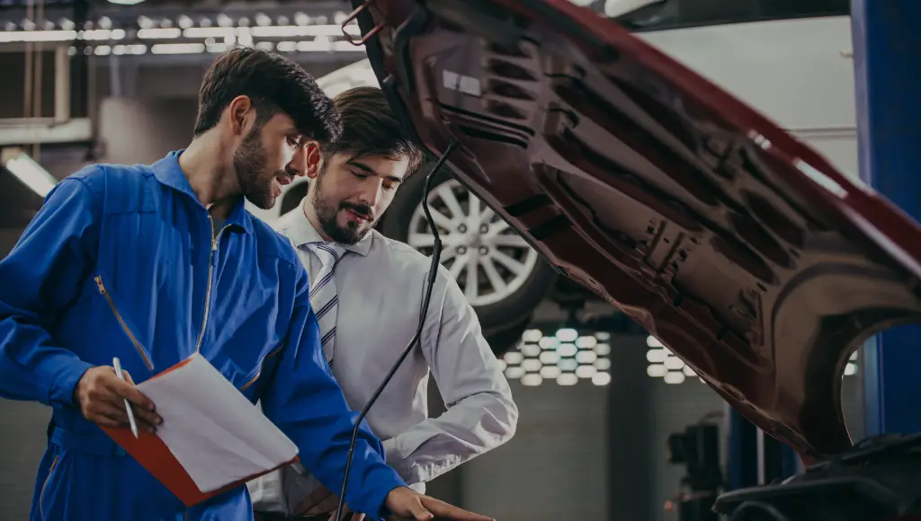 A mechanic and a man in a suit look under a car hood in an auto repair shop, discussing paperwork.