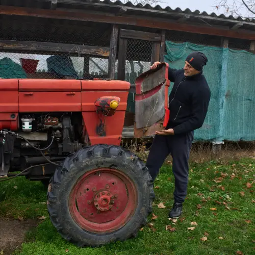 A man stands by a red tractor, holding its detached front panel near a shed on a grassy area.