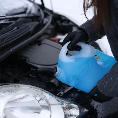 Person in gloves pouring blue windshield washer fluid into a car engine compartment on a snowy day.