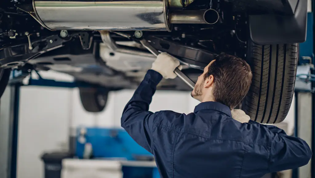 Mechanic in gloves working under a raised car in an auto repair shop, using a wrench for maintenance.