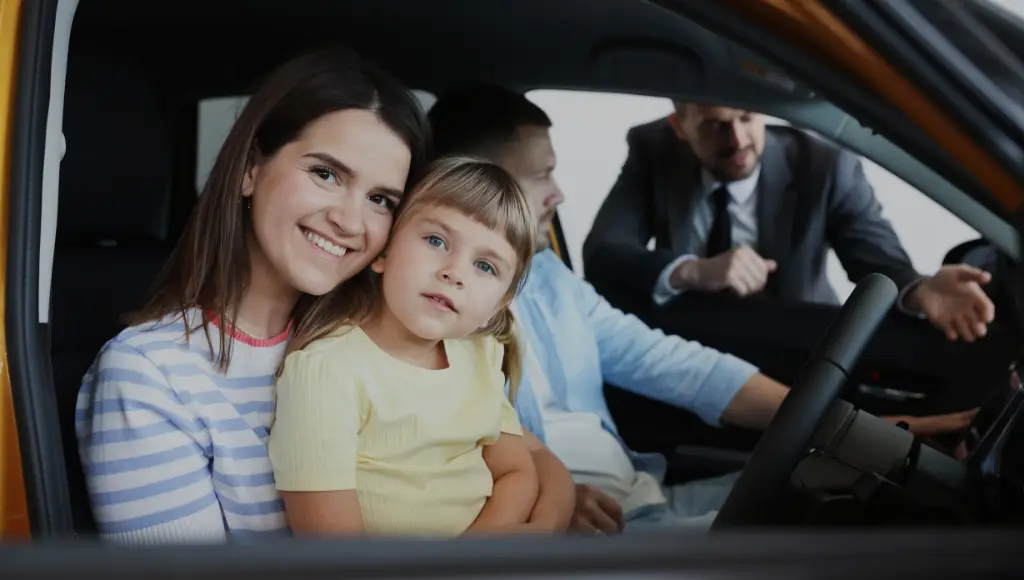 A smiling woman and child sit in a car, with two men talking outside the drivers window.