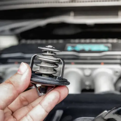 A hand holding a car thermostat in front of an open car engine compartment.