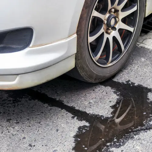 White car with a visible side front wheel parked over a puddle leaking fluid onto the asphalt.