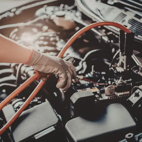 A gloved hand jump-starts a car battery with jumper cables under an open car hood.