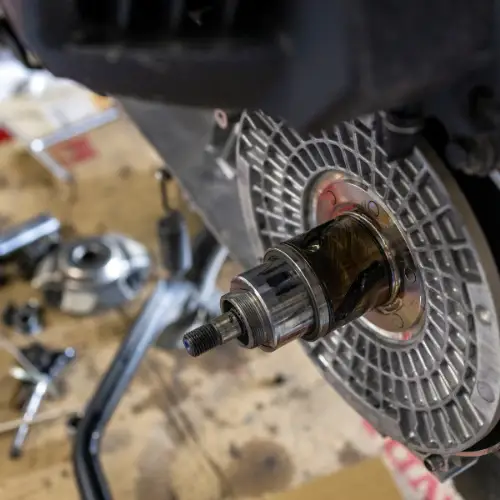 Close-up of a mechanical component, possibly a motorcycle engine part, with tools and parts blurred in the background.