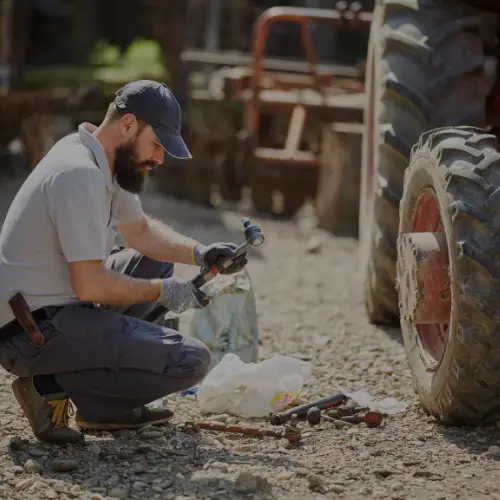 A man kneels by a tractor wheel, repairing equipment with tools scattered on the ground.