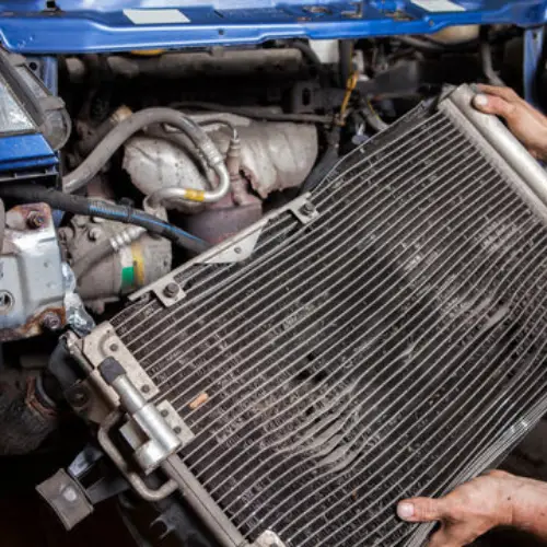 Mechanic’s hands removing a dirty car radiator from the engine bay of a blue vehicle.